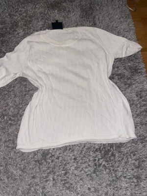 Vit ribbad t-shirt - En enkel vit t-shirt med ribbad struktur och rund hals. T-shirten har korta ärmar och är tillverkad i ett mjukt material som känns skönt mot huden. Perfekt till jeans eller shorts för en clean och avslappnad look.
