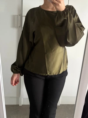 Olivgrön satinblus med ballongärm - Snygg olivgrön blus i glansigt satintyg med rund halsringning och långa ballongärmar med resår vid handlederna. Blusen har en avslappnad passform och ger en elegant look. Perfekt att styla med både jeans och kjol för en trendig outfit.