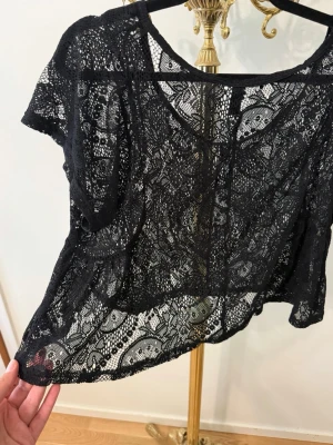 Svart spetsig croppad topp - Säljer en svart croppad topp i genombruten spets med korta ärmar. Toppen har ett detaljerat spetsmönster och är perfekt för att styla lager på lager. Den har en loose passform och rund halsringning.