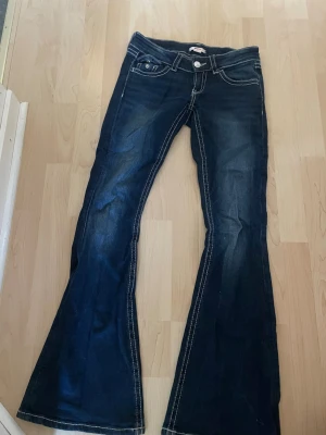 Mörkblå bootcut jeans med brodyr - Säljer ett par mörkblå bootcut jeans med vita kontrastsömmar och snygg brodyr på bakfickorna. Jeansen har låg midja, klassisk femficksdesign och utsvängda ben. Perfekta för dig som gillar en retrostil och vill sticka ut lite extra.
