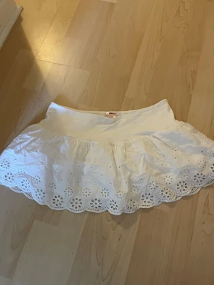 Vit kort kjol med broderade detaljer - Söt vit kort kjol med broderade blommor och vågig kant. Kjolen har en bred midja och är gjord i bomull, vilket gör den både luftig och bekväm. Perfekt för varma dagar och ger en fräsch och somrig look.