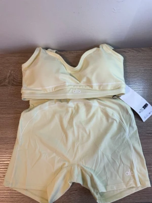Ljusgult träningsset från Alo Yoga - Fräscht ljusgult träningsset från Alo Yoga med sport-bh och matchande shorts. Setet har en clean design med smala axelband och elastisk midja. Tillverkat i mjukt, stretchigt funktionsmaterial som andas. Alo-logga syns framtill på både bh och shorts. Perfekt för yoga, gym eller dans.