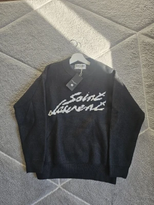 Svart stickad tröja Saint Laurent - Snygg mörkgrå stickad tröja från Saint Laurent med vit logotyp i handskriven stil på bröstet. Tröjan har rund hals, långa ärmar och ribbade muddar vid ärmslut och nederkant. Perfekt för dig som gillar stilrena och exklusiva plagg. Size S. Oanvänd köpte nyligen.