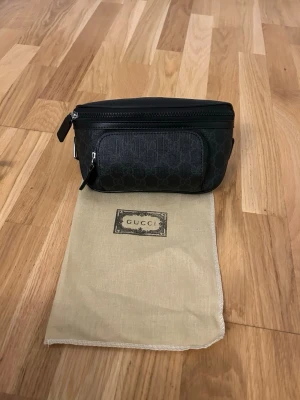 Svart Gucci bältesväska - Snygg svart bältesväska från Gucci i canvas med diskret GG-mönster. Väskan har två dragkedjefack, justerbart band med spänne och kommer med original dustbag. Perfekt för att bära det viktigaste på ett stilrent sätt.