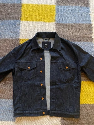 Mörkblå klassisk jeansjacka - Snygg mörkblå jeansjacka BOSS med raka linjer och klassisk krage. Jackan har två bröstfickor med lock och knappar i kopparfärg. Perfekt för dig som gillar en tidlös och stilren look. Långärmad och i robust denim.