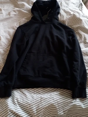 Svart hoodie med magficka - En klassisk svart hoodie med stor magficka och huva. Hoodien har ribbade muddar vid ärmslut och nederkant. Perfekt för en avslappnad och stilren look. Materialet känns mjukt och bekvämt, troligen bomullsblandning.