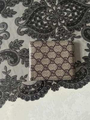 Beige och brun mönstrad plånbok - Snygg plånbok i beige och brunt med ett klassiskt monogrammönster. Utsidan har ett stilrent tryck medan insidan är svart med flera kortfack och ett fönster för ID-kort. Tillverkad i tåligt konstläder som känns lyxigt och modernt.