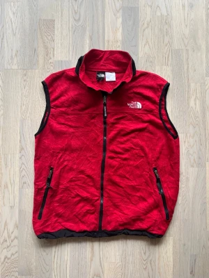 The North Face fleeceväst - L - Mycket bra skick. Inga tecken på användning Fleeceväst från The North Face, storlek L, röd Dragkedja med “The North Face”-text, två fickor med dragkedja, justerbar midja, röd med svarta detaljer Bröstvidd: 54 cm Längd: 64 cm Polyester, nylon Inga defekter ETIKETT BORTKLIPPT