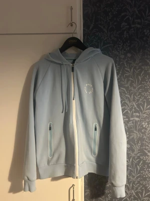 Ljusblå hoodie med dragkedja - Snygg ljusblå hoodie med dragkedja framtill och två fickor med dragkedja. Tröjan har huva med snören och ett diskret tryck på bröstet där det står 'Sosso for sosso'. Perfekt för en avslappnad och trendig stil.