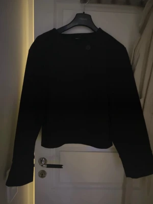 Svart kavaj med enkel knäppning - Stilren svart kavaj med rak passform och en enkel knapp framtill. Kavajen har rundad halsringning och långa ärmar. Perfekt för en clean och modern look. Materialet ser ut att vara mjukt och något tjockare, vilket ger en snygg siluett.