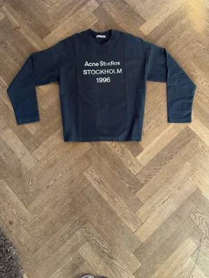 Svart t-shirt Acne Studios 1996 - Cool svart t-shirt från Acne Studios med vit text 'Acne Studios STOCKHOLM 1996' på bröstet. Klassisk rund halsringning och rak passform. Perfekt för dig som gillar stilrena och trendiga plagg med en touch av streetwear.