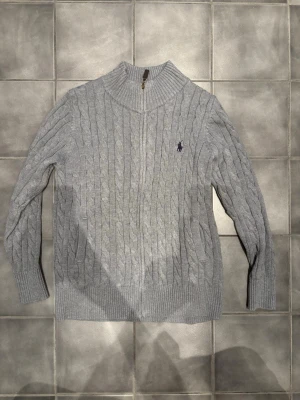 Grå kabelstickad tröja Polo Ralph Lauren - Kabelstickad grå tröja från Polo Ralph Lauren med halv dragkedja och hög krage. Tröjan har långa ärmar, ribbade muddar och den klassiska broderade loggan på bröstet. Perfekt för lager på lager och riktigt snygg till jeans.