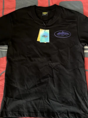 Svart t-shirt med lila tryck - Svart t-shirt i bomull med rund hals och korta ärmar. På bröstet finns ett lila tryck med motiv av en båt i en oval. Perfekt för dig som gillar stilrena plagg med en twist.