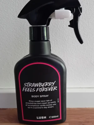 LUSH Strawberry Feels Forever Body Spray - En svart sprayflaska med rosa detaljer från LUSH, doften heter Strawberry Feels Forever. Kroppssprayen har en söt och fruktig doft av jordgubbar och vanilj. Flaskan rymmer 200 ml och har en praktisk sprayfunktion för enkel applicering.