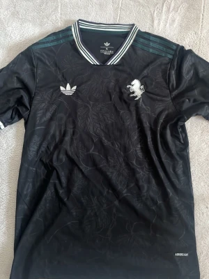 Svart Adidas  tröja - Snygg svart sporttröja. Tröjan har diskret bladmönster, vita Adidas-loggan -lejonet på bröstet samt klassiska Adidas-ränder på axlarna. Tillverkad i funktionsmaterial med AEROREADY-teknologi.