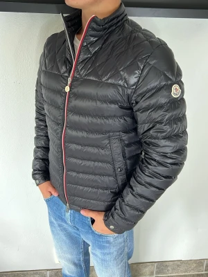 Moncler jacka - Snygg svart dunjacka från Moncler med quiltad design och hög krage. Jackan har dragkedja med rödvit kant, två sidofickor med knapp och Moncler-logga på ärmen. Perfekt för kalla vinterdagar och ger en stilren look.
