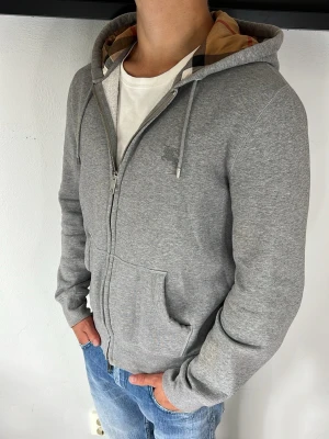 Burberry hoodie  - Snygg grå hoodie med dragkedja och huva. Insidan av huvan har ett rutigt mönster i beige och svart. Hoodien har fickor framtill och dragsko i huvan. Perfekt till jeans och enkel att matcha med andra plagg.