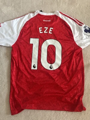 Arsenal matchtröja röd/vit nr 10 - Snygg Arsenal matchtröja i rött och vitt med nummer 10 och namnet EZE på ryggen. Tröjan har klubbmärke, Adidas-logga och Premier League-märke på ärmen. Tillverkad i lätt och ventilerande material, perfekt för fotbollsträning eller match.