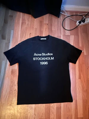 Svart Acne Studios t-shirt - Säljer en svart t-shirt från Acne Studios med vit text 'Acne Studios STOCKHOLM 1996' på bröstet. Klassisk rund hals och korta ärmar. Perfekt för dig som gillar stilrena och ikoniska plagg,säljer pga gilllar inte riktigt passformen och kan ej lämna tillbaka för jag har andvänt den 