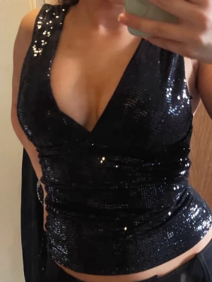 Svart glittrig topp - Snygg svart topp med paljetter och djup v-ringning. Toppen är ärmlös och har en figurnära passform som framhäver siluetten. Perfekt för dig som vill sticka ut med lite extra glitter. Nån paljett har lossnat nånstans men inget man tänker på
