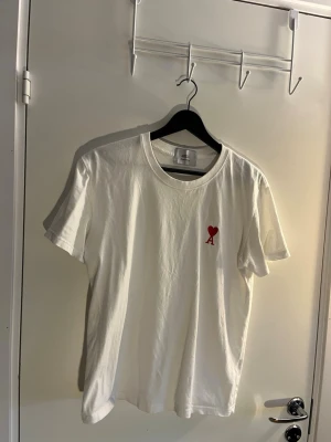 Vit t-shirt från Ami Paris - Säljer en clean vit t-shirt från Ami Paris med det klassiska röda hjärtat och A-loggan broderad på bröstet. T-shirten har rund hals och korta ärmar, perfekt basic-plagg i garderoben. Tillverkad i mjuk bomull för skön känsla.