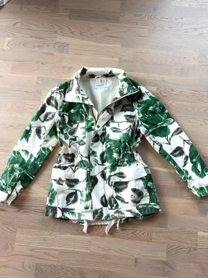 Grönblommig parkajacka från Ganni - Snygg parkajacka från Ganni med grönt blommönster på vit botten. Jackan har stora fickor framtill, dragsko i midjan och nederkant samt klassisk krage. Perfekt för dig som vill sticka ut med en unik och trendig jacka.
