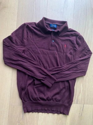 Vinröd stickad pikétröja Polo Ralph Lauren - Vinröd långärmad stickad pikétröja från Polo Ralph Lauren i mjuk merinoull. Klassisk krage med två knappar och broderad logga på bröstet. Perfekt för dig som vill ha en stilren och bekväm tröja med snygg passform. Tvättlapp av klippt tyvärr