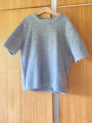Ljusgrå stickad t-shirt - Mysig ljusgrå stickad t-shirt med korta ärmar och rund hals. Tröjan har en avslappnad passform och ribbstickade detaljer vid ärmslut, halsringning och nederkant. Perfekt för lager på lager och enkel att styla till olika outfits.