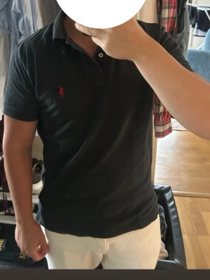 Svart pikétröja från Polo Ralph Lauren - Klassisk svart pikétröja från Polo Ralph Lauren med röd broderad logga på bröstet. Tröjan har korta ärmar, krage och knappar vid halsen. Tillverkad i mjuk bomull för en bekväm känsla och stilren look.