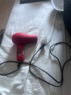 Röd hårfön med sladd - En kompakt och smidig hårfön i röd plast med svart sladd och tvåpolig kontakt. Perfekt för att snabbt torka håret och enkel att ta med på resan tack vare sin mindre storlek och lätta vikt.