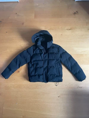 Svart replay pufferjacka med huva - Snygg svart Replay pufferjacka med huva och dragkedja framtill. Jackan har två framfickor och quiltad design som ger en modern look. Perfekt för kyliga dagar och enkel att matcha med olika outfits.