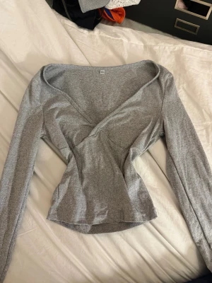 Grå omlottopp med v-ringning - Säljer en stilren grå omlottopp med v-ringning och långa ärmar. Toppen har en figurnära passform och är tillverkad i mjukt bomullsmaterial. Perfekt att matcha med jeans eller kjol för en enkel men snygg look.
