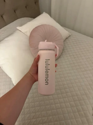 Ljusrosa vattenflaska Lululemon - Snygg ljusrosa vattenflaska från Lululemon med praktiskt lock och handtag. Flaskan är tillverkad i tåligt material och har en stilren design med Lululemon-loggan tryckt vertikalt på sidan. Perfekt att ha med till gymmet, skolan eller på språng. Helt ny och oanvänd!!