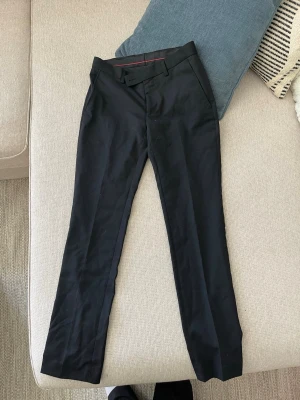 Svarta kostymbyxor Dressmann - Snygga svarta kostymbyxor från Dressmann med raka ben och klassisk passform. Byxorna har bälteshällor, sidofickor och är tillverkade i ett mjukt, vävt material som känns bekvämt hela dagen. Perfekta för en clean och stilren look.