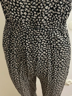 Svartvit blommig jumpsuit - Säljer en svart jumpsuit med små vita blommor i ett tätt mönster. Modellen har resår i midjan för en skön passform och långa ben. Materialet ser ut att vara mjukt och följsamt, perfekt för en avslappnad look.