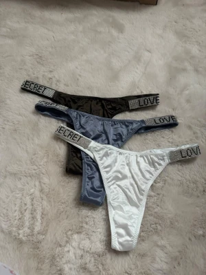 Satinstring med strass och textdetalj - Tre snygga stringtrosor i satinmaterial i färgerna svart, blå och vit. Trosorna har breda resårband dekorerade med glittrande strass och texten 'LOVE SECRET' i svart. Perfekt för dig som gillar glam och trendiga detaljer.