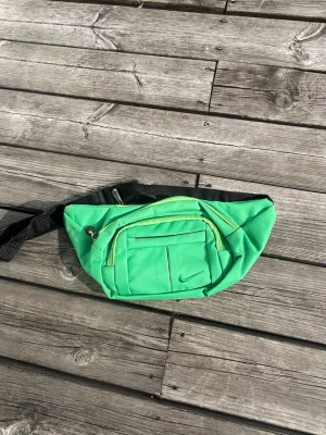 Grön magväska från Nike - Snygg grön magväska från Nike med svart justerbart band och flera fack med dragkedja. Perfekt för att bära det viktigaste när du är på språng. Klassisk Nike-logga på framsidan och praktisk design.