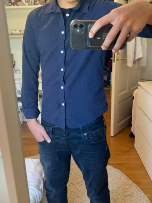 Mörkblå Tommy hilfigher skjorta - Snygg mörkblå skjorta med klassisk krage och vita knappar framtill. Skjortan har lång ärm och en liten diskret logga på bröstet. Perfekt till jeans eller chinos för en clean och stilren look.