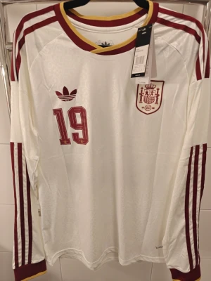 Vit långärmad Spanien tröja Adidas - Säljer en vit långärmad fotbollströja från Adidas med Spaniens landslagsemblem och nummer 19 i vinrött. Tröjan har vinröda ränder längs ärmarna och gul detalj vid kragen. Perfekt för dig som gillar retrostil och fotboll.