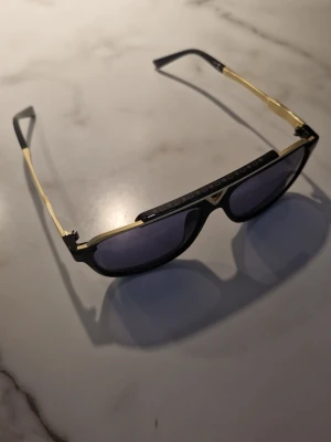 Louis Vuitton svarta solglasögon med gulddetaljer - Snygga solglasögon från Louis Vuitton med svart båge och guldfärgade detaljer på sidorna och överdelen. Glasen är mörka med  lätt lila ton och har LV-loggan ingraverad. Modell Mascot kantig och modern form som ger en lyxig vibe. Självklart äkta lite använda. Louis Vuitton:↙️ https://en.louisvuitton.com/eng-nl/men/accessories/sunglasses/gold/black/_/N-t194fv1y-axbx9vr3_pb5r44?page=3.                                           Nypris: 6250:-. Kvitto skickas när betalningen är helt genomförd.