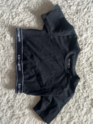Svart croppad topp från Carhartt - Säljer en svart croppad topp från Carhartt med korta ärmar och elastiskt band nertill med Carhartt-logga. Toppen är i mjuk bomull och har en enkel, sportig look som passar perfekt till jeans eller kjol.