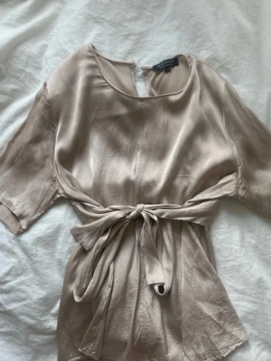 Champagnefärgad omlottopp med knyt - Snygg champagnefärgad topp i silkesliknande material med korta ärmar och rund hals. Toppen har ett knytband i midjan som ger en omlottkänsla och en feminin siluett. Perfekt att styla med jeans eller kjol för en trendig look.