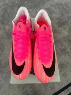 Nike rosa mercurial med svart swoosh - Säljer ett par grymma Nike löparskor i neonrosa med stor svart swoosh på ovansidan. Skorna har snörning, vit insida och en futuristisk genomskinlig sula med gröna detaljer och Zoom-teknologi. Perfekta för dig som vill sticka ut på löprundan!