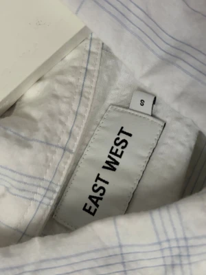 Vit rutig skjorta från EAST WEST - Klassisk vit skjorta från EAST WEST i storlek S med tunna blå rutor. Skjortan har lång ärm, bröstficka och knappar framtill. Perfekt för en clean och stilren look. Tillverkad i bomull för en skön känsla.