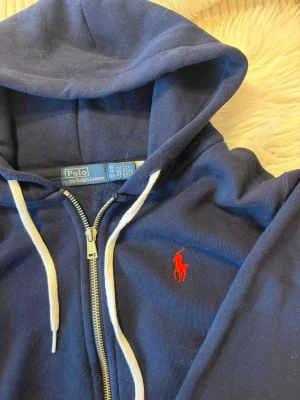 Marinblå hoodie från Polo Ralph Lauren - Snygg marinblå hoodie med dragkedja från Polo Ralph Lauren. Klassisk design med vit dragsko i huvan och röd broderad logga på bröstet. Tillverkad i mjukt bomullsmaterial som känns skönt mot huden. Perfekt för en avslappnad och stilren look.