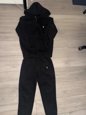 Svarta mjukisbyxor+ hoodie från Polo Ralph Lauren - Säljer ett par svarta mjukisbyxor+ hoodie från Polo Ralph Lauren med den klassiska vita loggan broderad på benet. Byxorna har dragsko i midjan och är gjorda i mjukt bomullsmaterial som är riktigt skönt att ha på sig. Perfekta för chill dagar eller när du vill vara bekväm.