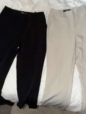 Två par stilrena byxor i svart och beige/vit - Säljer två par byxor, ett par svarta och ett par beige/vit. De svarta har slitsar vid bensluten och en smal passform, medan de beige är raka och klassiska. Perfekta för en clean och enkel look. Materialet ser ut att vara mjukt och stretchigt, troligen polyesterblandning.
