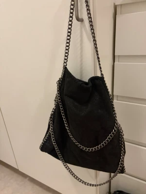 Svart handväska med kedjedetaljer - Cool svart handväska med silverfärgade kedjor som axelrem och runt kanterna. Väskan har en mjuk, lite slouchy form och dragskostängning upptill. Perfekt för dig som gillar en edgy och trendig look.