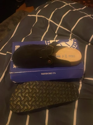 Svarta Birkenstock Boston tofflor - Säljer ett par svarta Birkenstock Boston tofflor i mocka med klassisk korksula och svart räfflad yttersula. Tofflorna har en justerbar rem med metallspänne och är perfekta för chill dagar hemma eller när du vill ha det bekvämt.