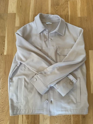 Beige overshirt med fickor - Stilren beige overshirt med klassisk krage och knäppning framtill. Två bröstfickor med lock och knappar samt två sidofickor. Overshirten har raka ärmar med manschett och är tillverkad i ett mjukt, strukturerat tyg som ger en avslappnad look. Helt ny, aldrig använd från zara
